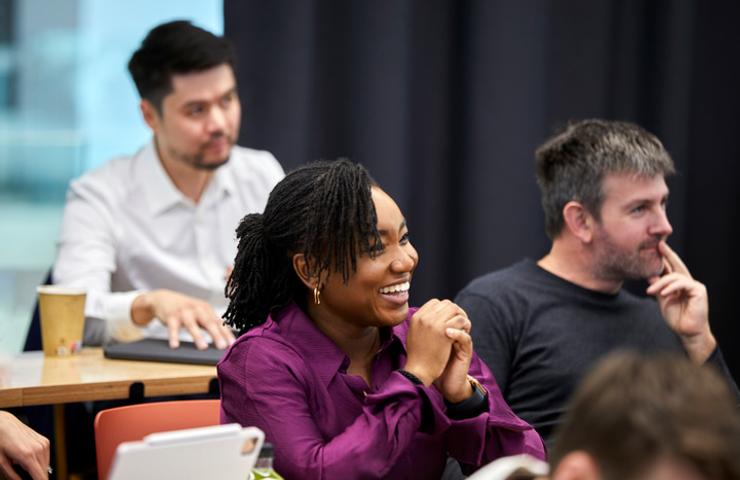 A group of people sitting in a meeting room, listening and interacting with each other, with a smiling woman in a purple blouse in focus.