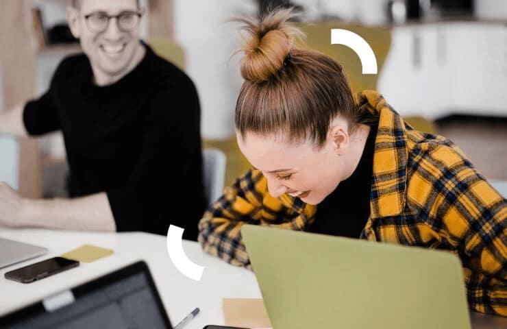 A woman in a plaid shirt works on a laptop with a smiling man in the background in a bright office environment.