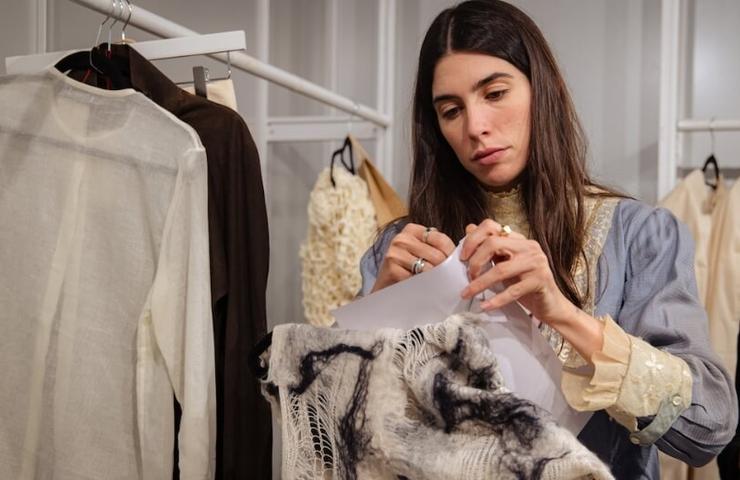 A woman examines a piece of fabric in a fashion design studio surrounded by garments on racks.