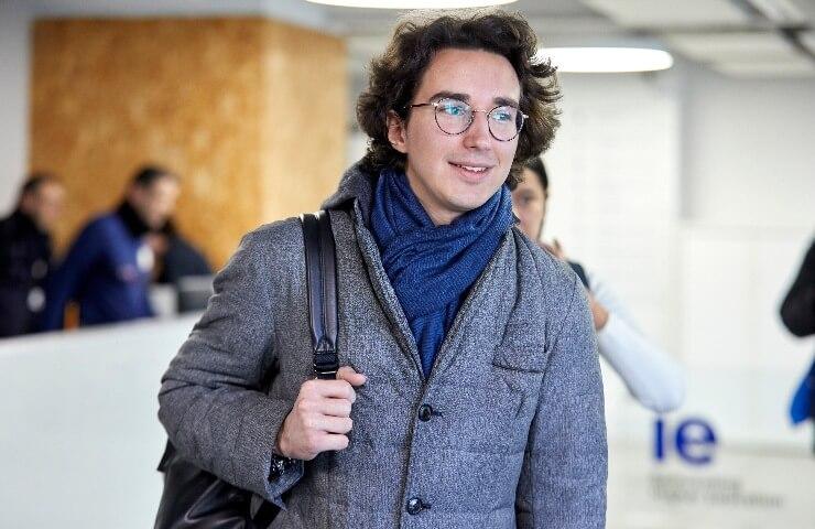 A young man wearing a coat and scarf carrying a backpack in a modern office environment.