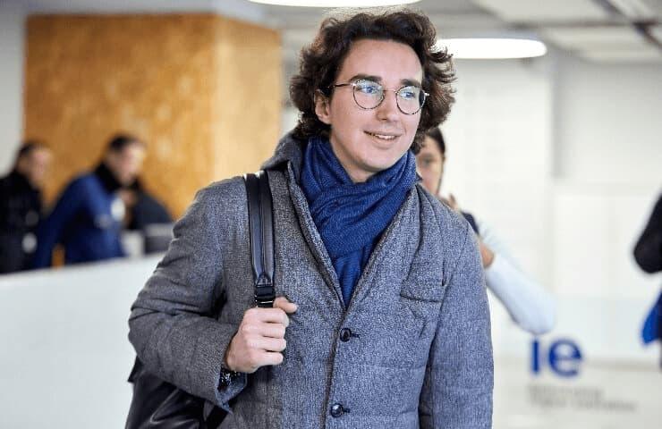 A young man with a backpack walking through an indoor area, wearing a grey coat and a blue scarf.