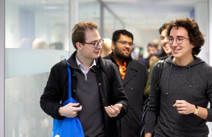 Group of four men walking and talking in an office corridor.