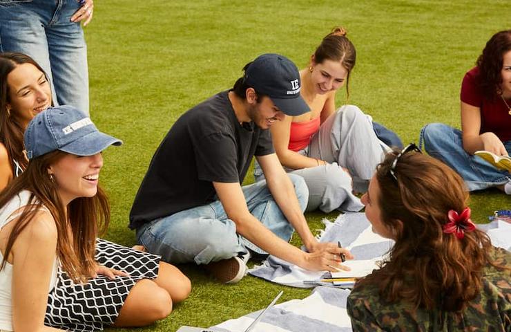 A group of young adults studying together outdoors on a grassy area with laptops and notebooks.