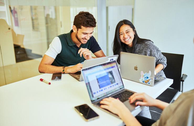 Two young adults are smiling and looking at a laptop screen in a modern office environment.