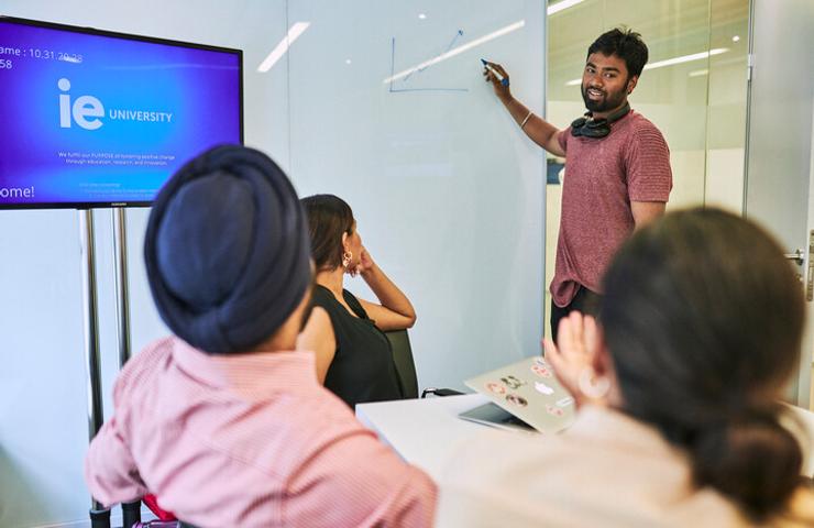 A man is presenting to a group of three people in a modern classroom with a digital screen displaying 'IE University' in the background.