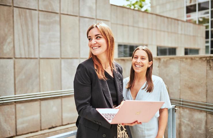 Two women smiling and holding a laptop outdoors in a city environment.