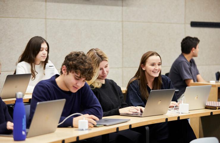 Students sitting at desks in a classroom, working on laptops and interacting with each other.