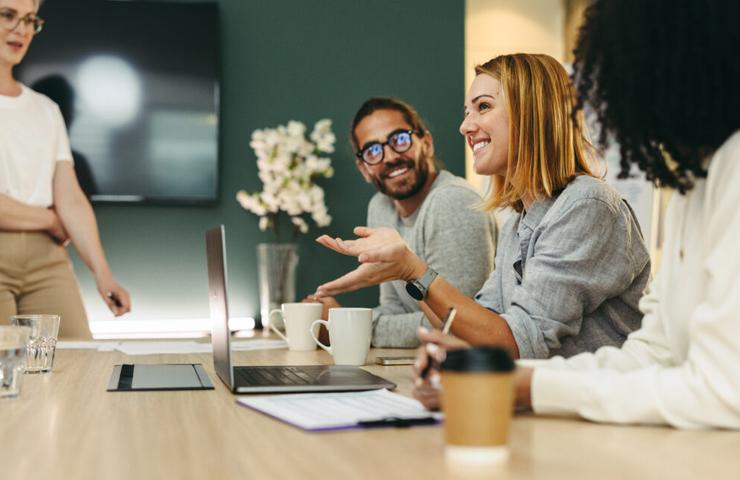 A group of diverse coworkers are engaged in a discussion around a table with laptops and coffee cups.