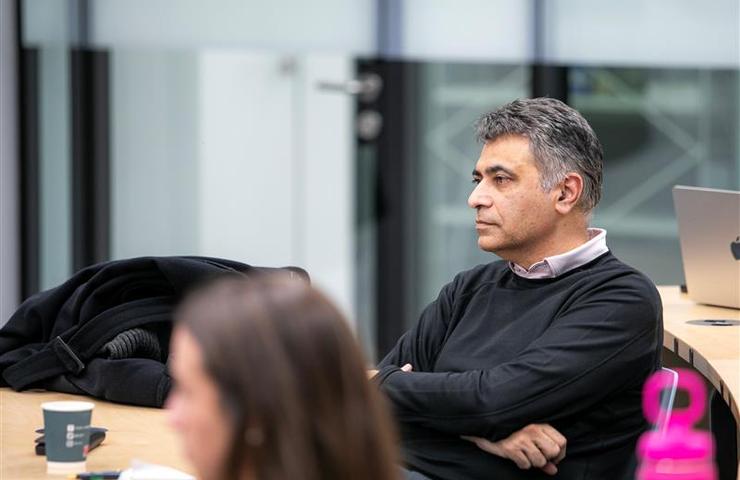 A man in a meeting room is seated attentively listening, while another person in foreground is out of focus.