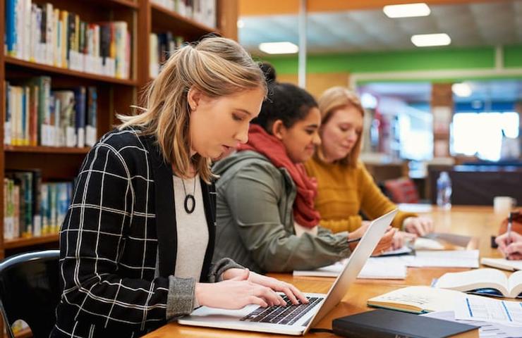 A young woman using a laptop in a library with fellow students studying around her.