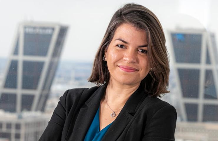 A professional woman with a confident smile sitting in an office environment, with skyscrapers visible through the window behind her.