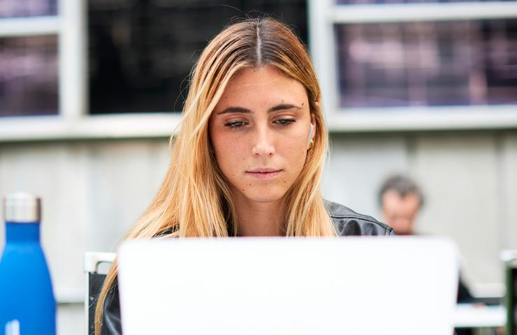 A young woman focusing intently on her laptop in an outdoor setting, possibly on a university campus.