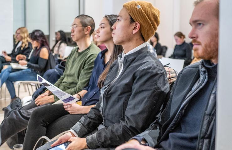 A group of attentive people seated in a row at an indoor event, focusing forward.