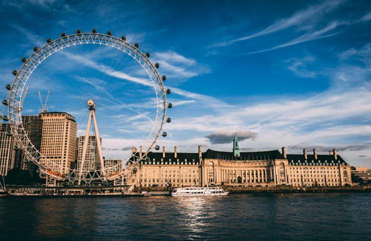 A view of the London Eye and surrounding buildings along the River Thames under a blue sky.