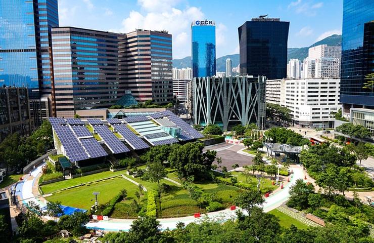 A modern urban park with solar panels and lush greenery surrounded by tall commercial buildings and a clear blue sky.