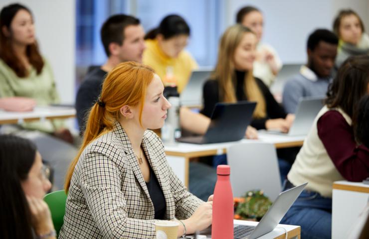 A diverse group of students attentively listening in a classroom setting, one of whom is a red-haired woman using a laptop.