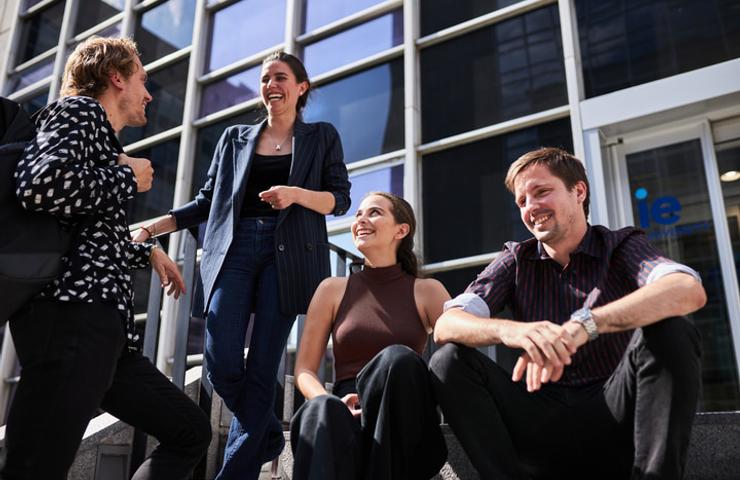 Four people happily chatting and laughing outside a modern office building on a sunny day.