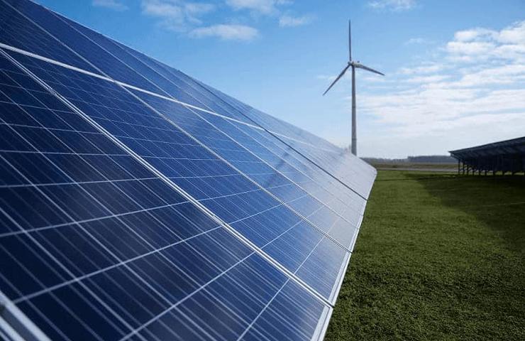 A solar panel array in the foreground with a wind turbine in the background under a clear sky.