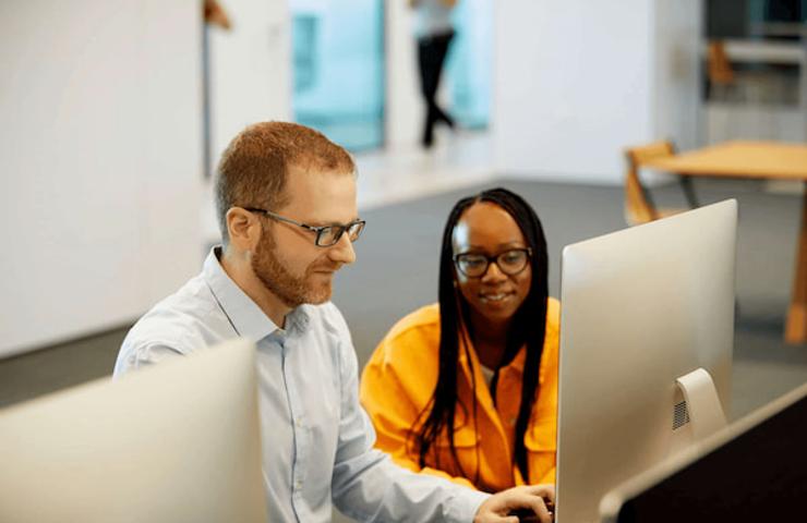 Two colleagues, a man and a woman, are working together at a computer in a modern office setting.