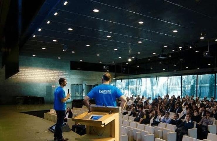 Two speakers in blue shirts presenting to an audience in a modern conference hall.