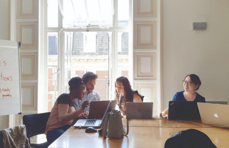 A diverse group of four professionals engaged in a discussion while sitting at a wooden table with laptops in a brightly lit room.