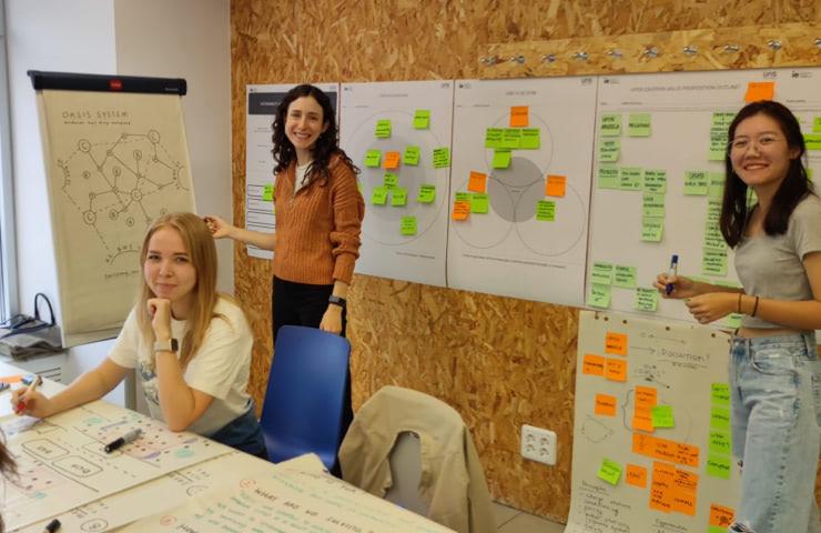 Three women smiling and working on project planning with sticky notes and charts on whiteboards in a creative workshop environment.