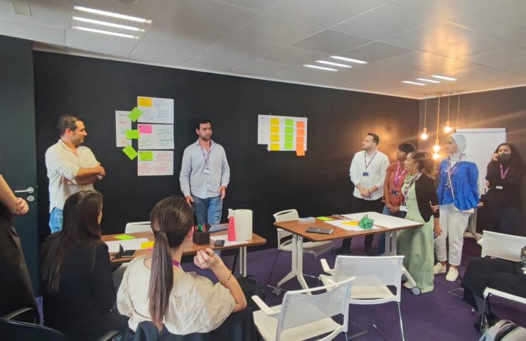 A group of professionals is engaged in a discussion in a modern office setting, surrounded by colorful notes and a whiteboard.