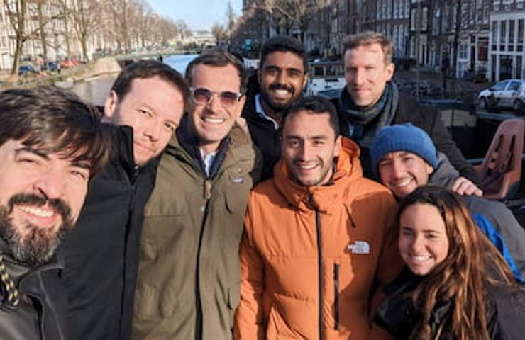 A group of seven people posing together for a selfie on a sunny day in a city with bicycles and canals visible in the background.