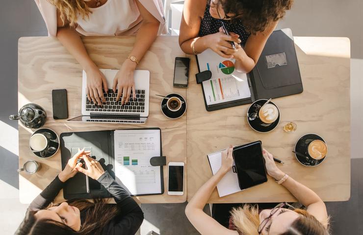 Top view of three individuals working around a table with laptops, notebooks, and coffee cups.