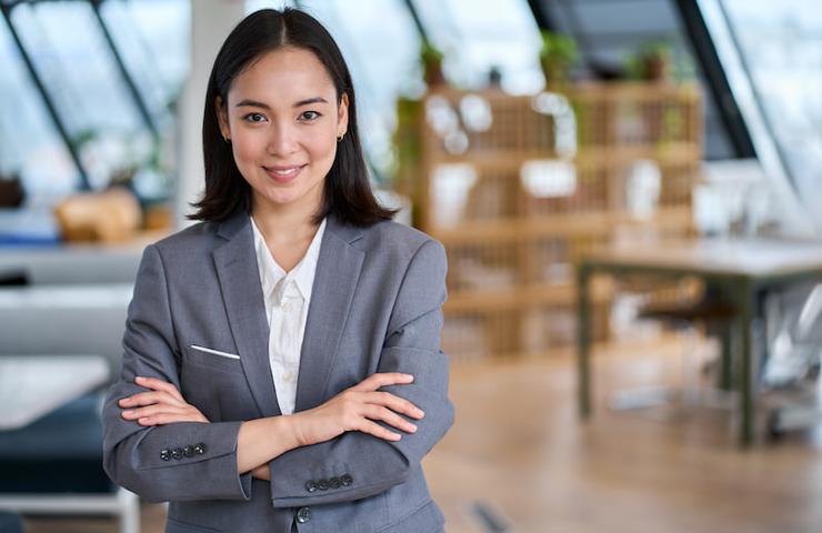 A confident businesswoman in a grey suit standing with her arms crossed in a modern office setting.