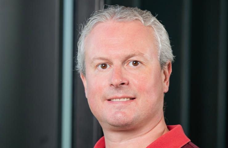 A professional portrait of a middle-aged man with light hair wearing a red shirt, standing in front of a window with blinds.