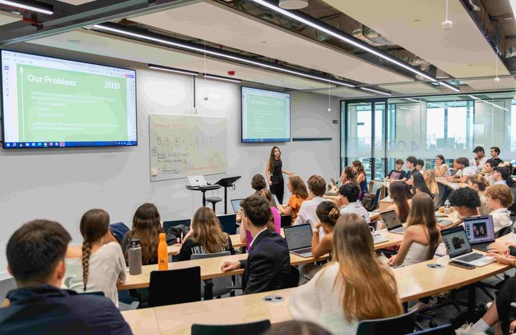 A person is presenting in a classroom full of students seated at desks with laptops.