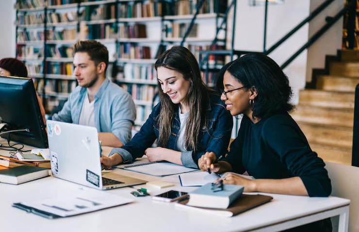 Three people, two women and one man, are sitting at a desk in a library, working on laptops and taking notes.