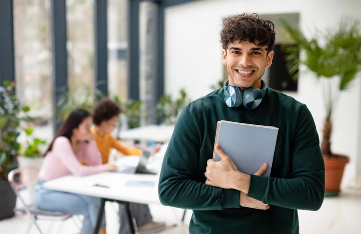 A young man with headphones around his neck holding a laptop smiles at the camera, with people in the background focusing on their work in a bright, modern workspace.