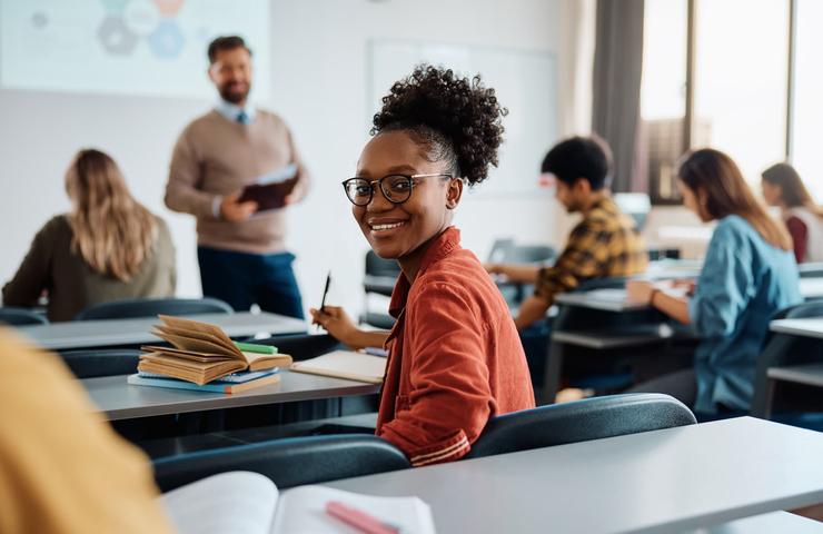 A young female student smiling at the camera while sitting in a classroom with peers and a teacher in the background.