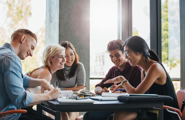 A group of five young adults happily studying together at a table in a sunny room.