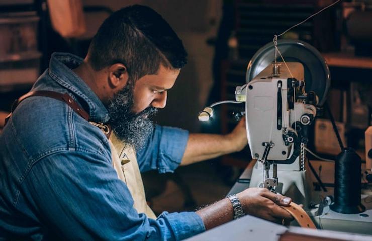 A man with a beard is focused on sewing fabric using an industrial sewing machine in a workshop.