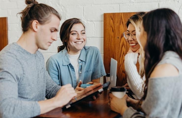 Four young adults are sitting at a table in a cafe, engaging in a joyful conversation while one of them uses a tablet.