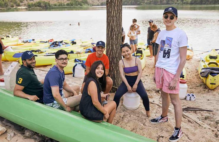 A group of young adults smiling and sitting by a lake with colorful kayaks in the background.