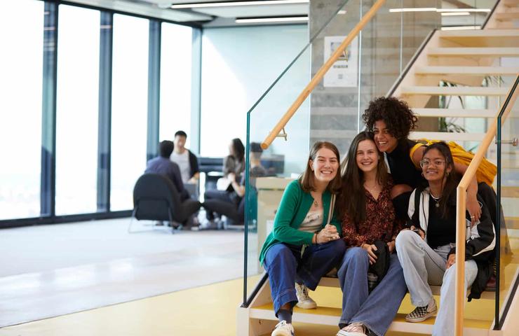 Four young adults smiling and sitting on yellow stairs in a modern indoor setting.