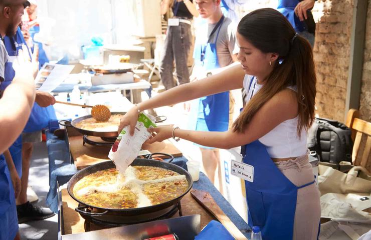 A woman in a blue apron is cooking in a large pan at a busy outdoor event.