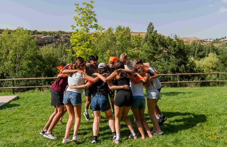 A group of people in a huddle outdoors in a grassy area with trees and a clear sky.