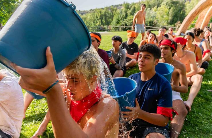 A young person is splashed with water from a blue bucket by another person in a sunny outdoor setting with a group of onlooking teenagers.