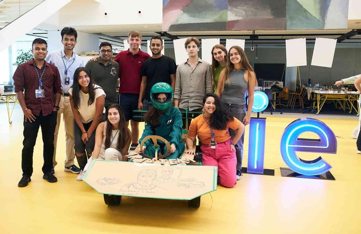 A group of diverse young adults gather around a table with a Google sign in the background at an apparent tech or educational event.