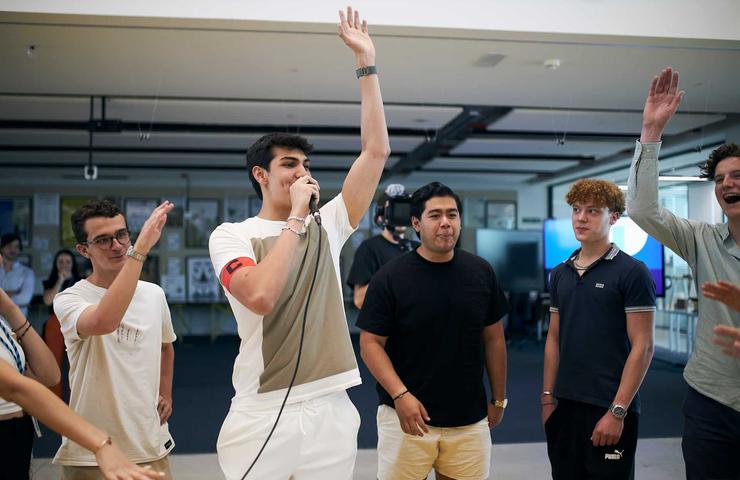 A young man enthusiastically singing into a microphone surrounded by cheering friends in a casual indoor setting.