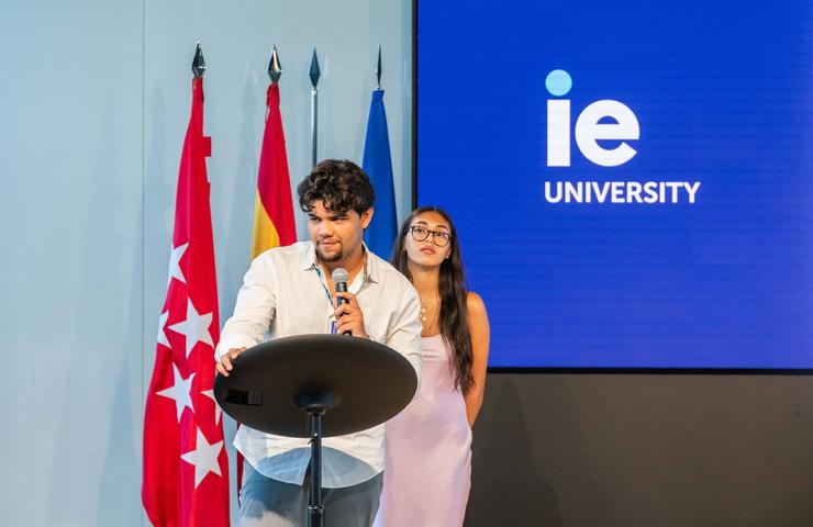 A young man is speaking at a podium with a microphone, while a young woman stands next to him at an event in a room with flags and a university's logo in the background.