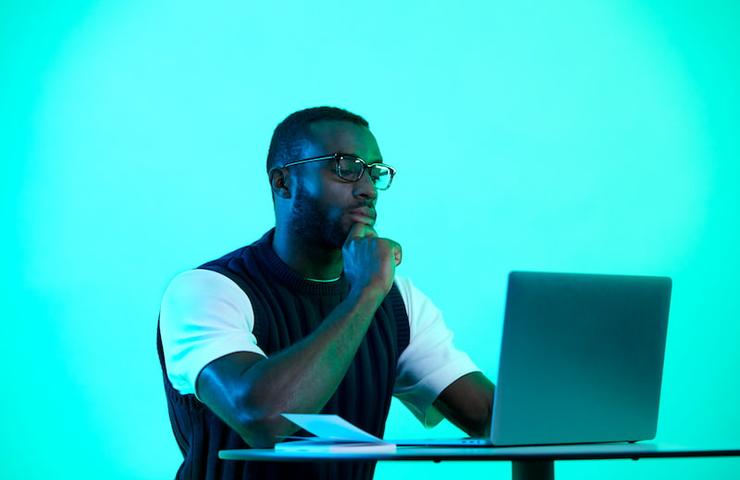 A man in glasses is sitting at a desk, deep in thought while looking at a laptop in a room with green lighting.