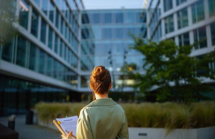 A woman standing outside, facing a modern building complex, holding papers and a yellow book.