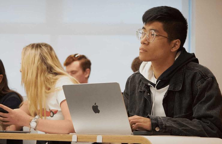 A young man with glasses and a denim jacket sits at a desk with a laptop in a classroom, looking to the side while other students are in the background.