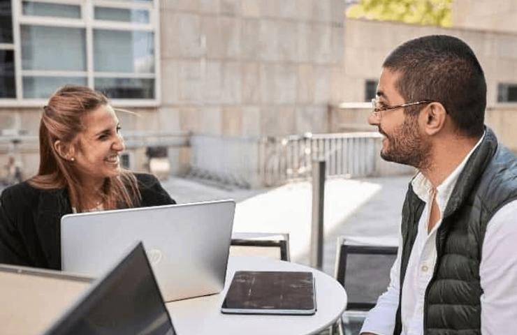 Two professionals smiling and engaging in a conversation at an outdoor table with a laptop and digital tablet.